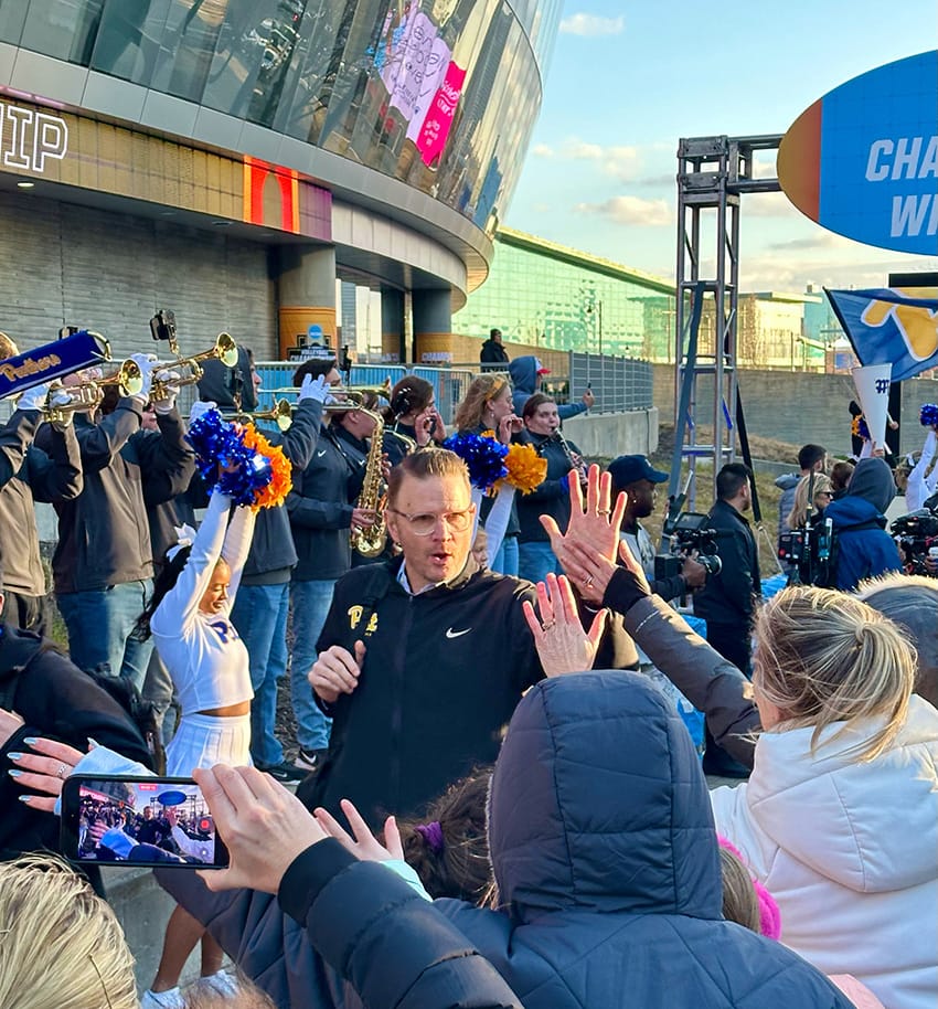 Photo of Pitt Volleyball head coach Dan Fisher high-fiving fans with the Pitt band in the background in Kansas City