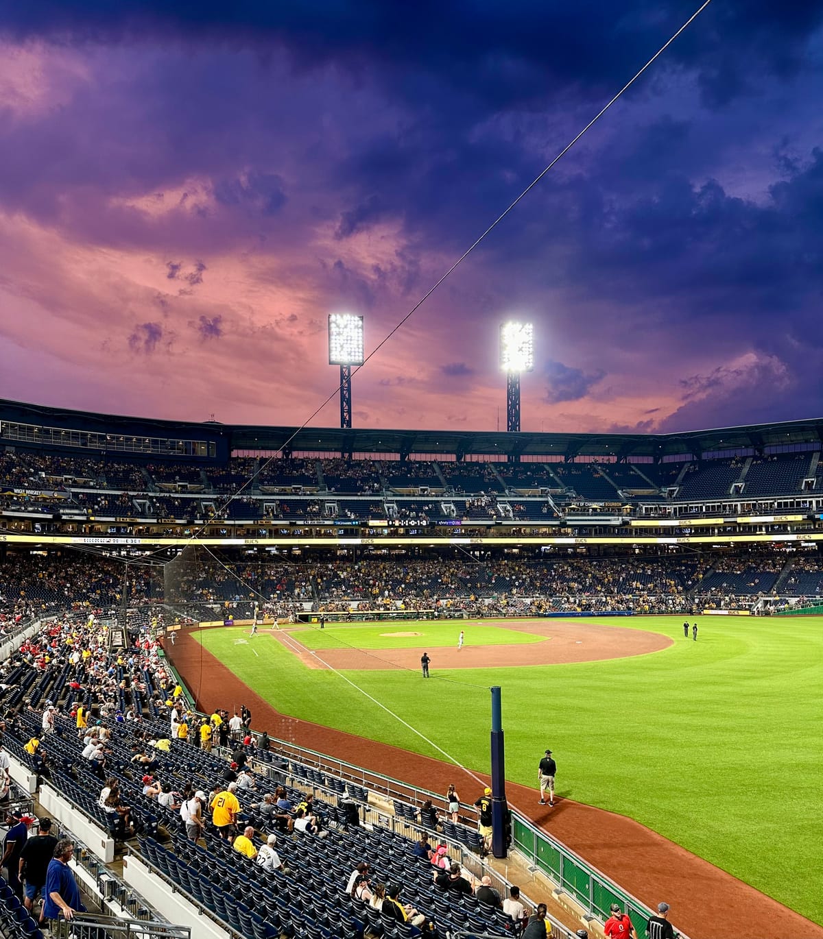 An evening photo of PNC Park in Pittsburgh from the right field seats. It shows the playing field, stands, lights, and a vibrant pink and purple sky at dusk