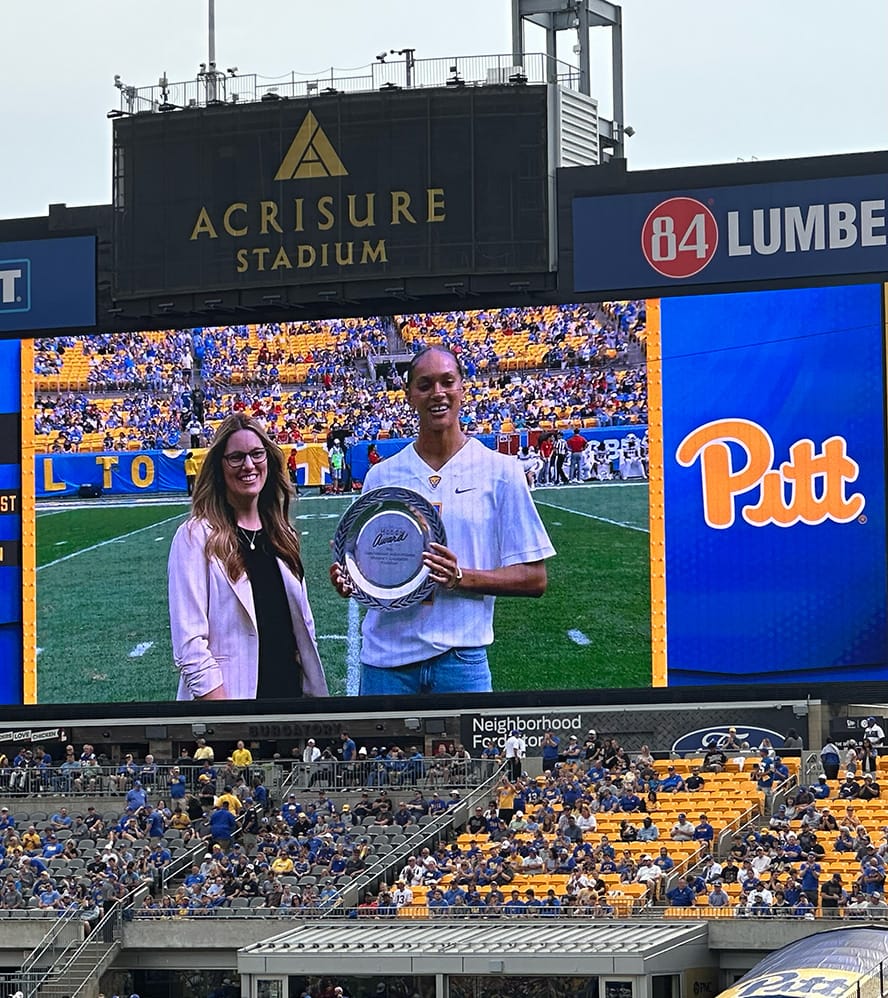 Okay, the photo shows Pitt Volleyball's Olivia Babcock holding a silver plate for the Honda Award for volleyball, seen on a stadium scoreboard